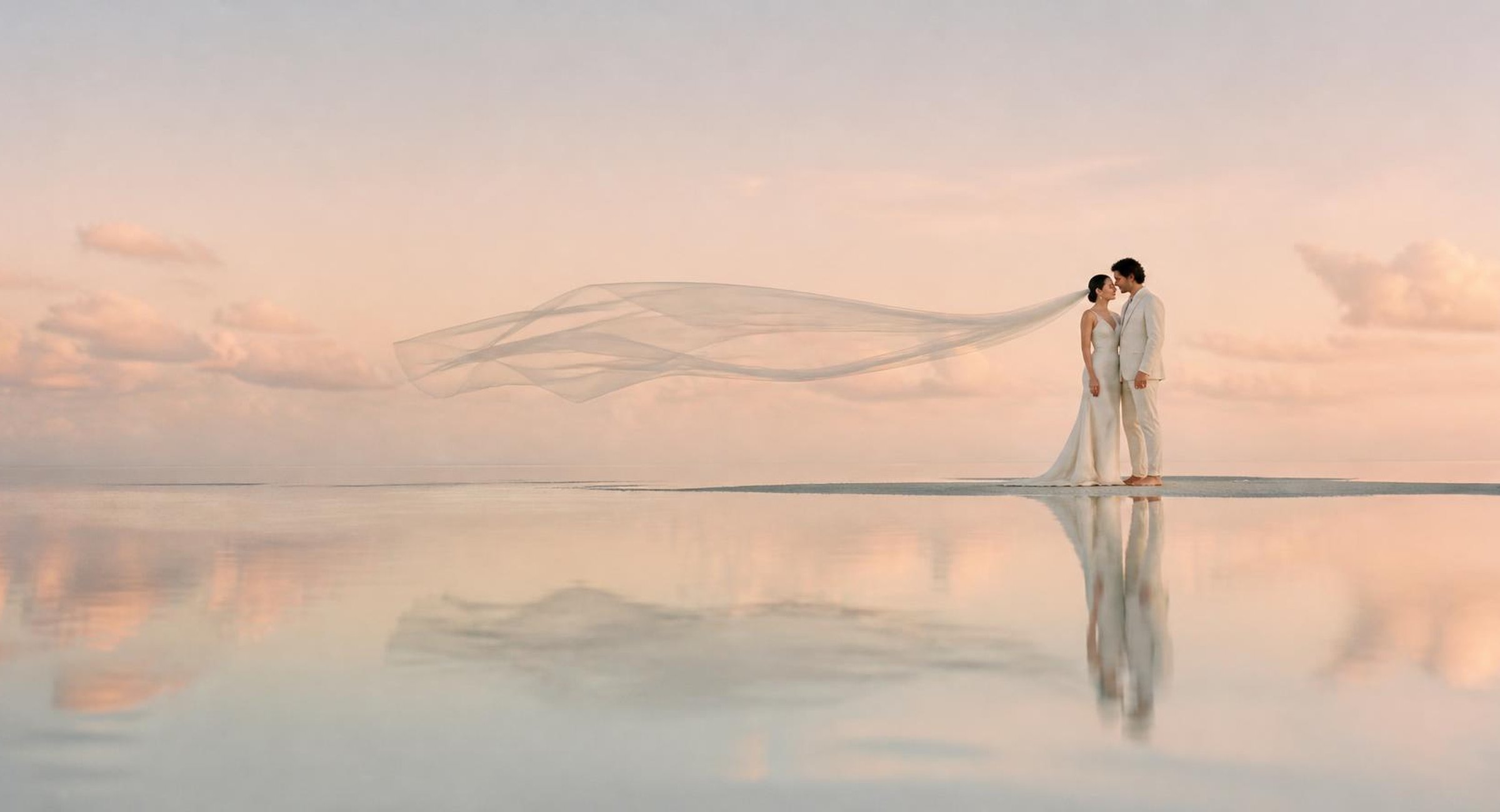 Bride and groom on a mirror-still sandbar at sunrise, her long veil streaming in the wind